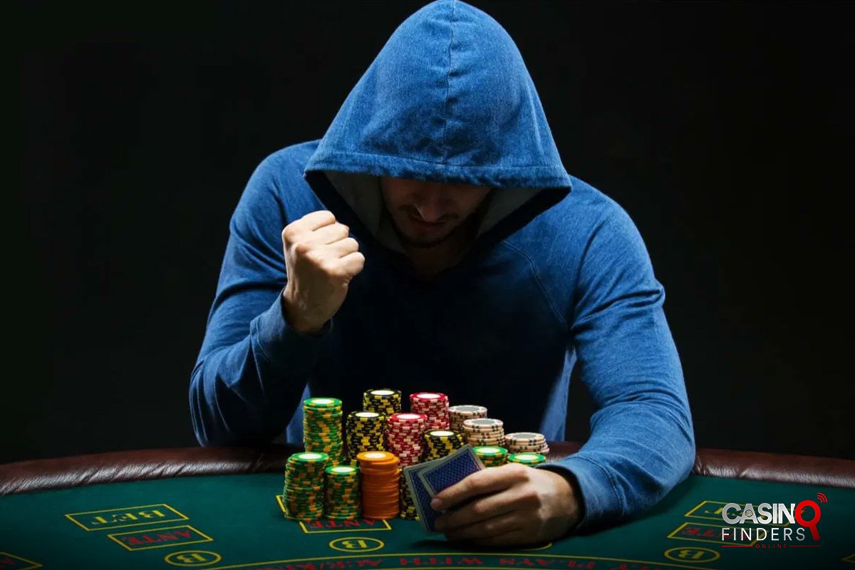 A male poker player wearing a navy blue hoodie sitting at a poker table holding playing cards in his hands